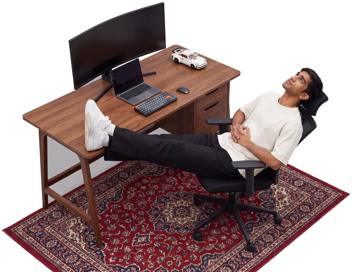 Mohit leaning back in an office chair, feet up on a walnut desk with a curved monitor, MacBook, mechanical keyboard and a Lego Porsche 911, on a red Persian rug, staring at the ceiling.